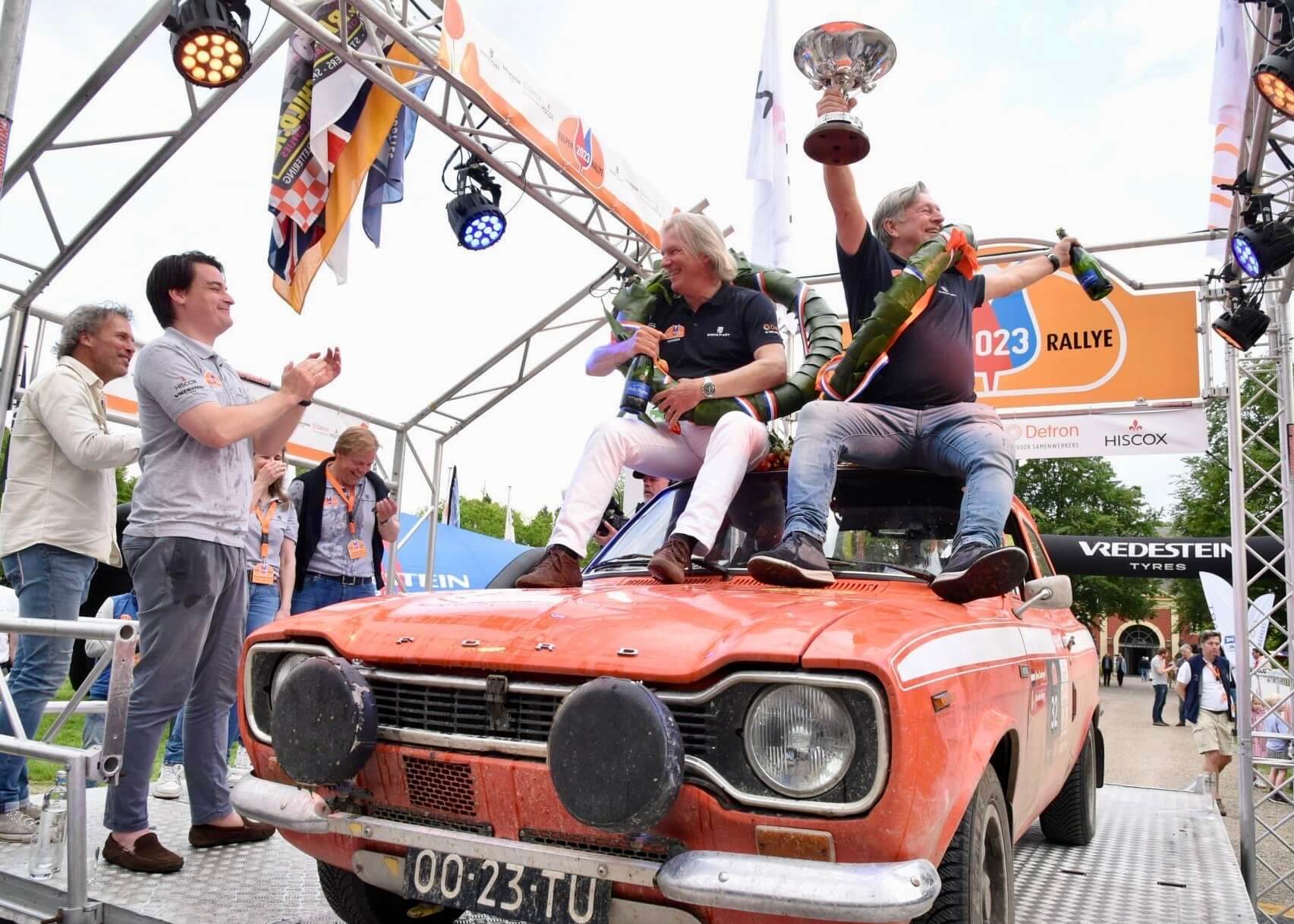 Winners of Tulip Rally celebrating on top of their car with trophy and champagne and supporters congratulating them
