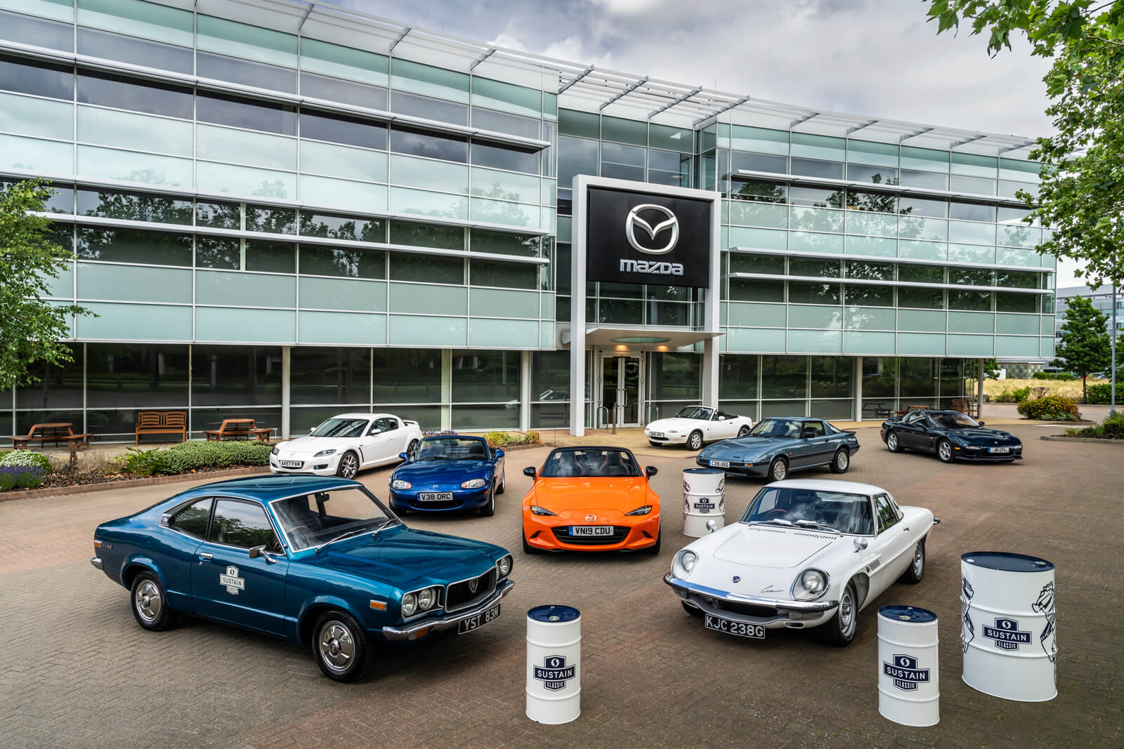 Vergrößerte Ansicht zahlreicher Fahrzeuge der Mazda UK Heritage Fleet, die ein Fass mit SUSTAIN Classic-Kraftstoff umgeben, vor einem Mazda-Gebäude